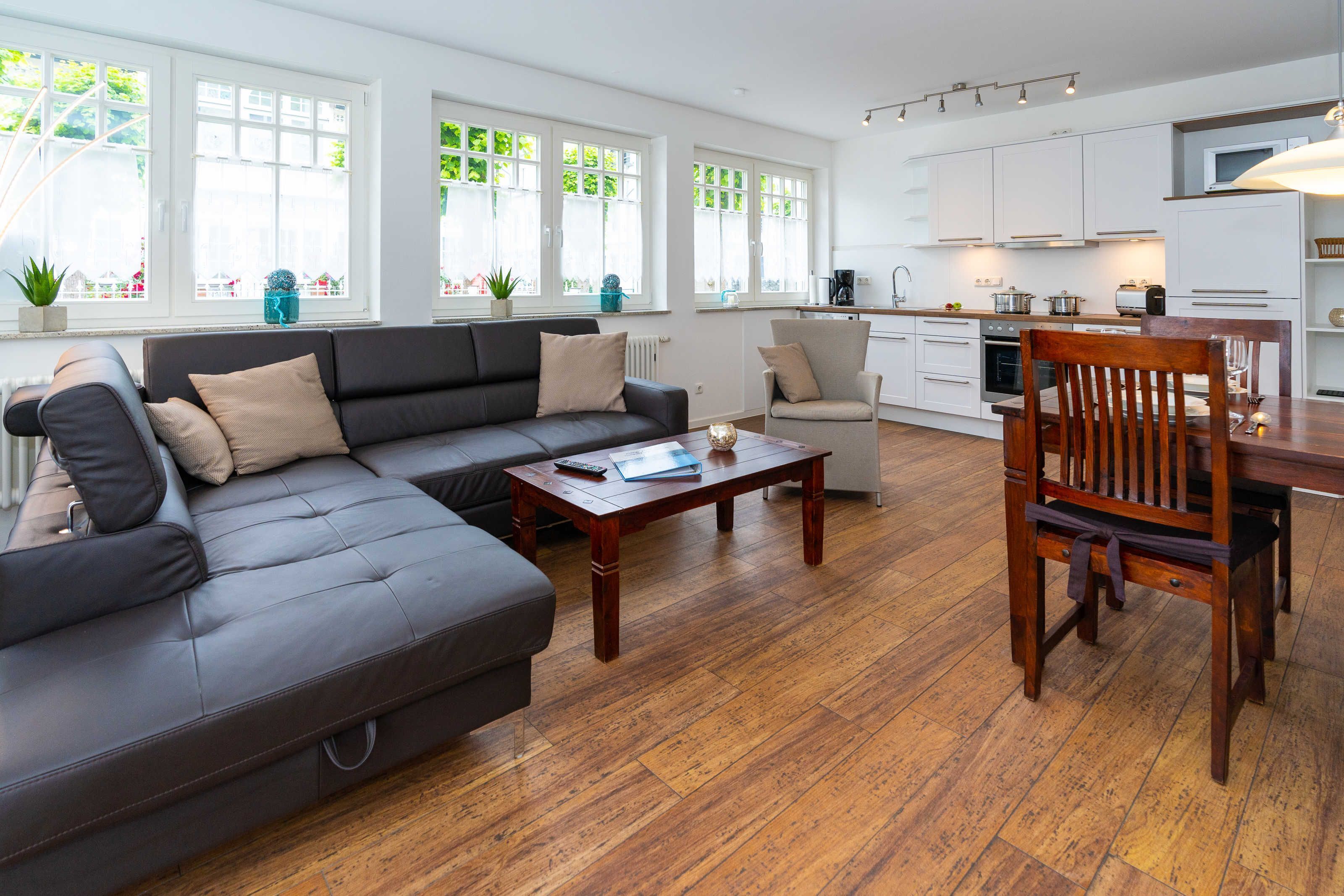 A modern living room with a sectional sofa, a wooden coffee table, and a dining area with a wooden table and chairs. There are large windows providing natural light and a view of greenery outside.