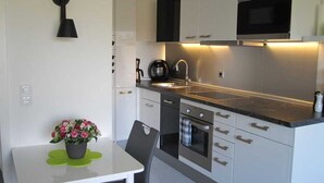 Et moderne køkken med hvide skabe, en sort bordplade, en indbygget ovn og en mikrobølgeovn. Der er et lille spisebord med en stol og en potteplante.