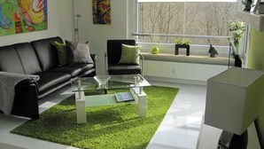 En moderne stue med en sort lædersofa, et glasbord og en grøn måtte. To billeder hænger på væggen, et vindue med udsigt til træer, og en radiator.