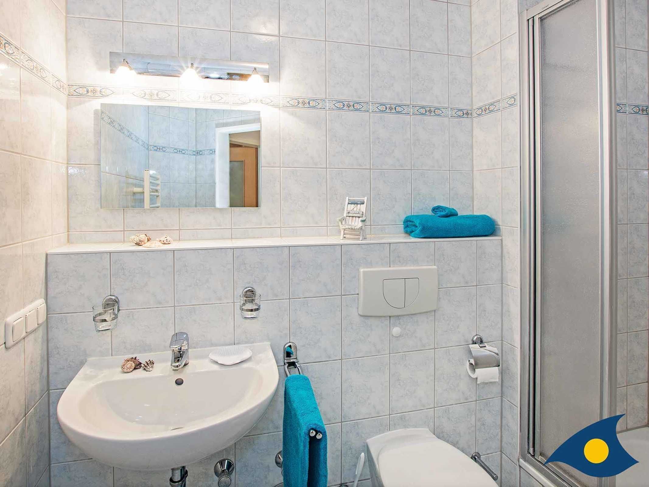 Une salle de bain équipée d’un lavabo, de toilettes et d’une douche. On y trouve un porte-serviettes avec une serviette bleue et une petite chaise.