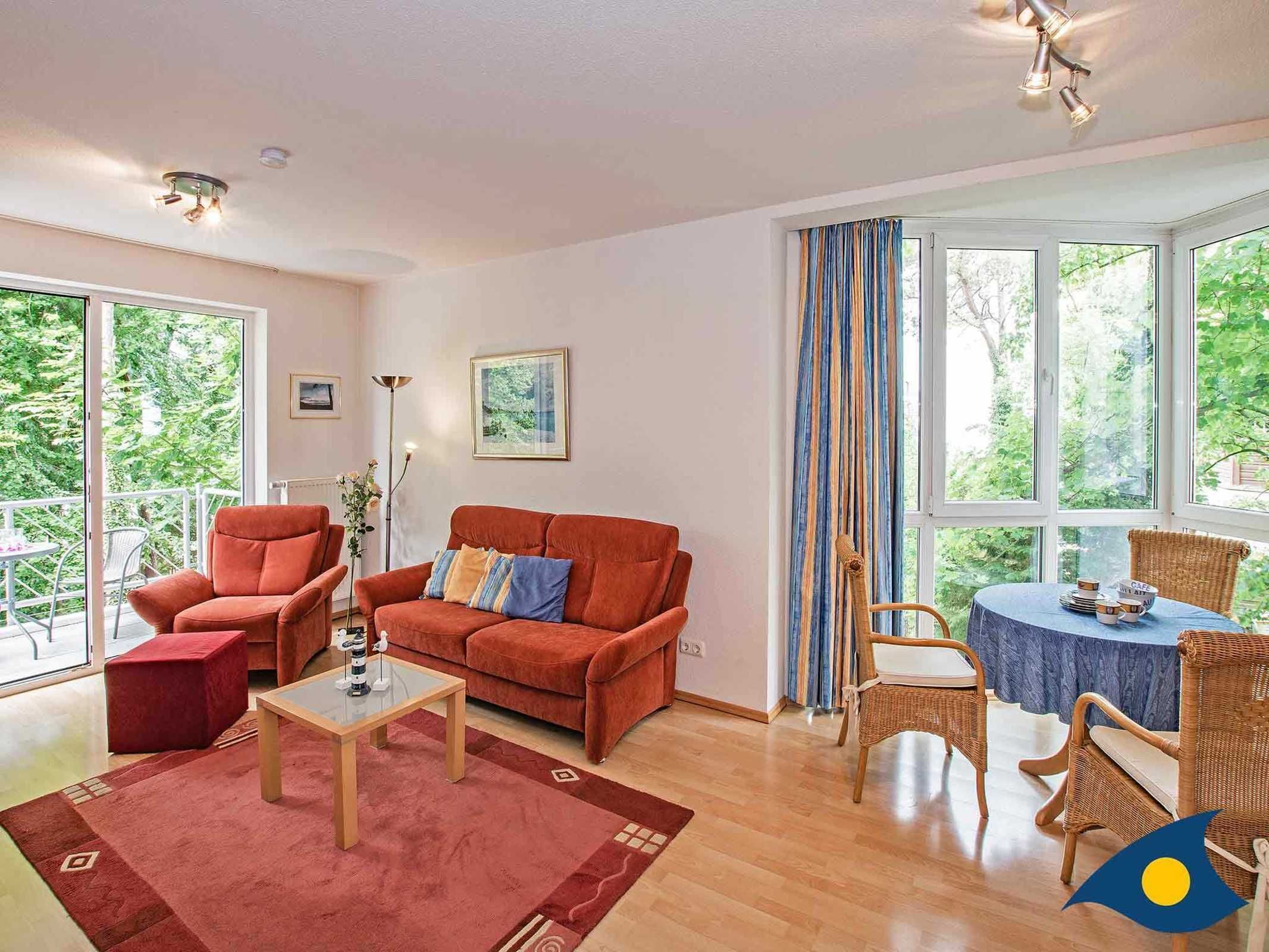 Un salón amplio con un sofá rojo, una mesa de centro de cristal y una mesa de mimbre con sillas. Grandes ventanas ofrecen vistas al verdor exterior.