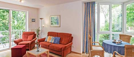 Un salón amplio con un sofá rojo, una mesa de centro de cristal y una mesa de mimbre con sillas. Grandes ventanas ofrecen vistas al verdor exterior.