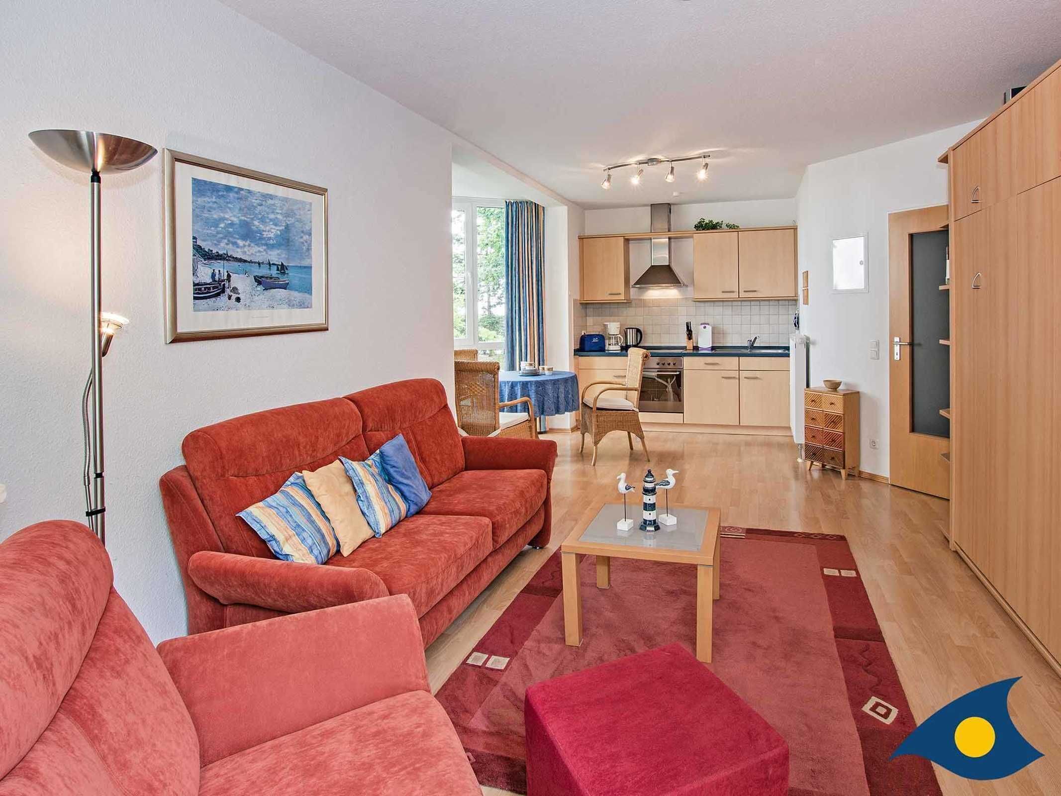 A modern living room with a red sofa, a coffee table, and a floor lamp. There is a kitchen in the background with wooden cabinets and a dining area.