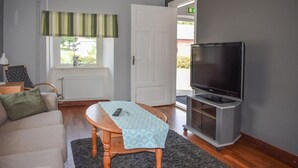 En stue med en sofa, et træbord med en fjernbetjening, et fjernsyn på et stativ, et vindue med en stribet gardin, og en dør der fører ud.