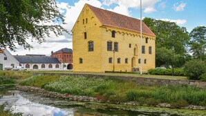 En gul bygning med rødt tag, en sø med siv og en stenvæg.