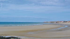 Ein weiter Sandstrand mit sanften Wellen, einem klaren Himmel und einer fernen Küstenlinie mit Gebäuden.