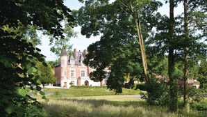 Ein großes, historisches Gebäude mit einem markanten Turm und symmetrischer Architektur, umgeben von üppiger Vegetation und einem gepflegten Rasen.