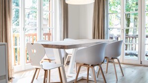 Ett modernt matområde med ett träbord och vita stolar, stora fönster med gardiner och en hängande lampa.