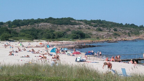 Una spiaggia con persone che prendono il sole e nuotano, tende e una collina rocciosa sullo sfondo.