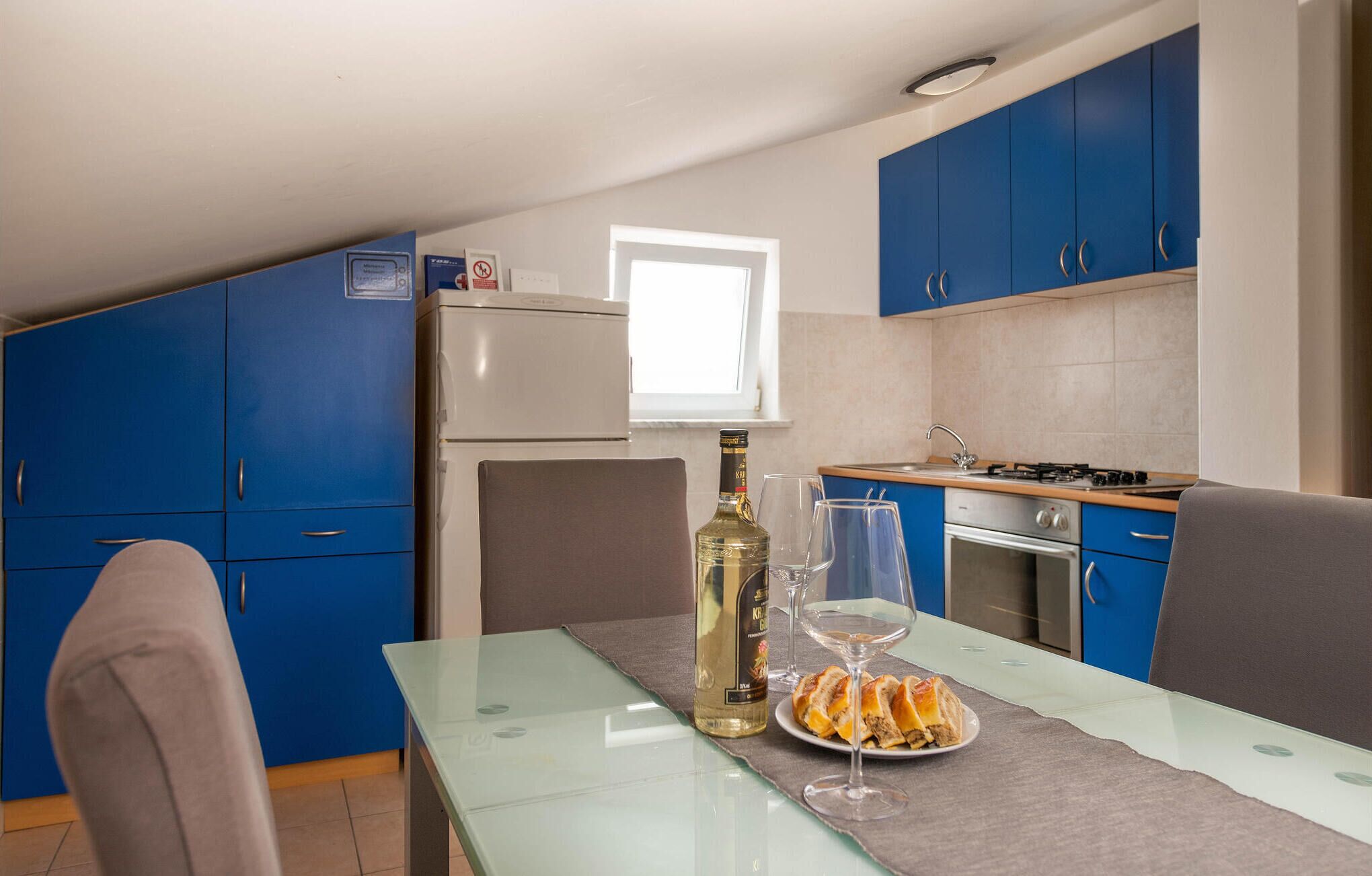 Een compacte keuken met blauwe kastjes, een witte koelkast, een gasfornuis en een kleine eettafel met een glazen blad.
