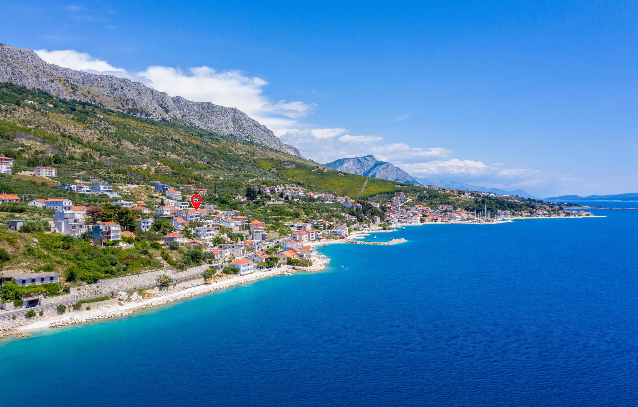 Un pueblo costero con edificios a lo largo de la costa, un mar azul cristalino y montañas al fondo.