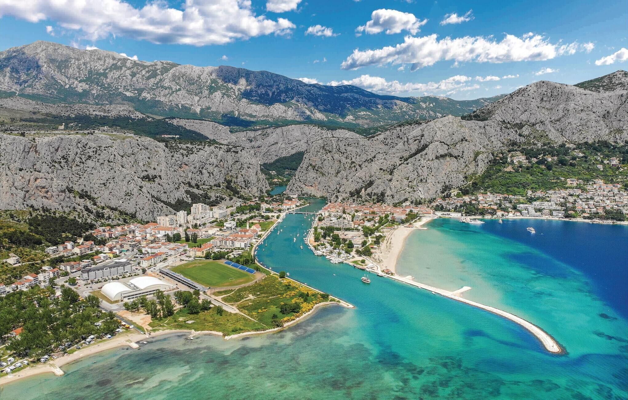 Luchtbeeld van een kustplaatsje met een jachthaven, helder turquoise water en een bergachtig landschap op de achtergrond.