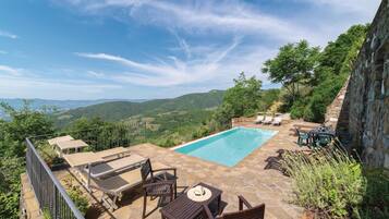 Area piscina con sedie a sdraio e un tavolo, circondata da muri in pietra e vegetazione, con vista su un paesaggio montuoso.