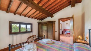 A bedroom with a wooden ceiling, a window, a wardrobe, a bed with a colorful bedspread, and a view of a dining area through an open door. - Nice apartment in Loro Ciuffenna AR (Loro Ciuffenna AR)