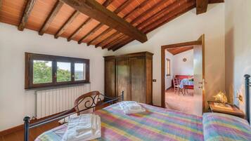 Una camera da letto con soffitto in legno, una finestra, un armadio, un letto con copripiumino colorato e vista su un'area pranzo attraverso una porta aperta.