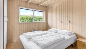 Quarto com teto de madeira, uma janela ampla com vista para o verde, uma cama com roupa de cama branca e uma mesa de cabeceira com abajur.