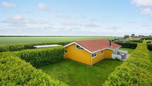 Um edifício amarelo com telhado vermelho, rodeado por sebes bem cuidadas e um campo verde ao fundo.