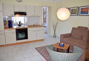 A modern kitchen with white cabinets, a built-in oven, and a dining area with a wicker chair and a small table.