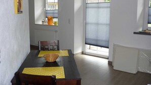 A dining area with a dark wooden table, yellow placemats, and a woven basket. There are two windows with blinds, a radiator, and a view of the outside. - Apartment 1 - Horn: Vacation in a thatched house in Schleswig-Holstein (Silberstedt)