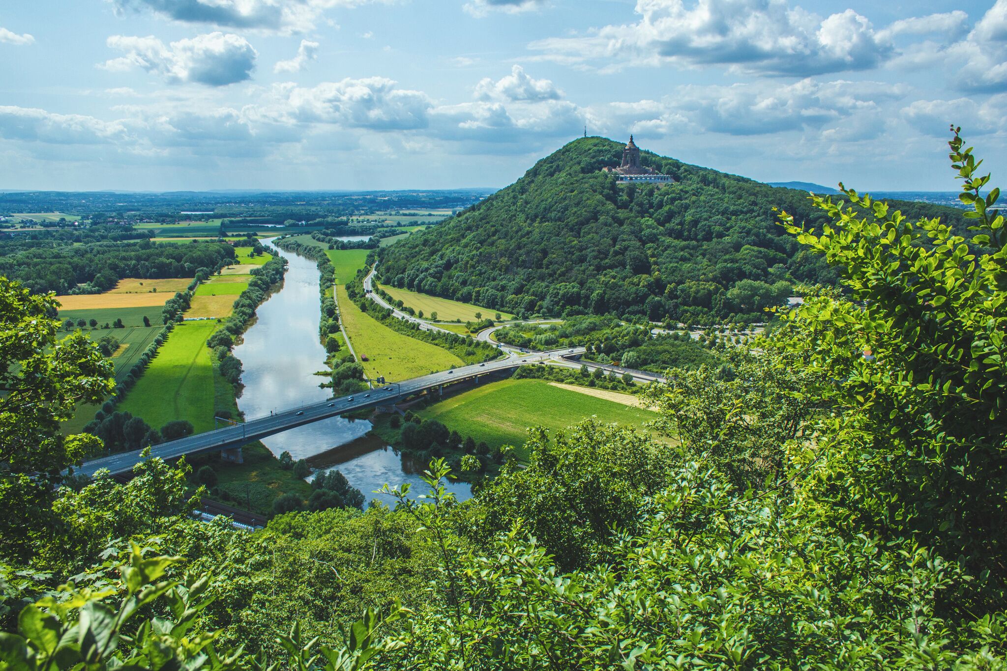 A lush green landscape with a river, a bridge, and a hill with a structure on top.