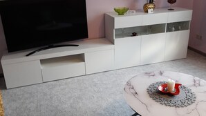 Ett vardagsrum med en platt-TV, ett vitt skåp, ett litet bord med ett ljus och en väggmonterad lampa.