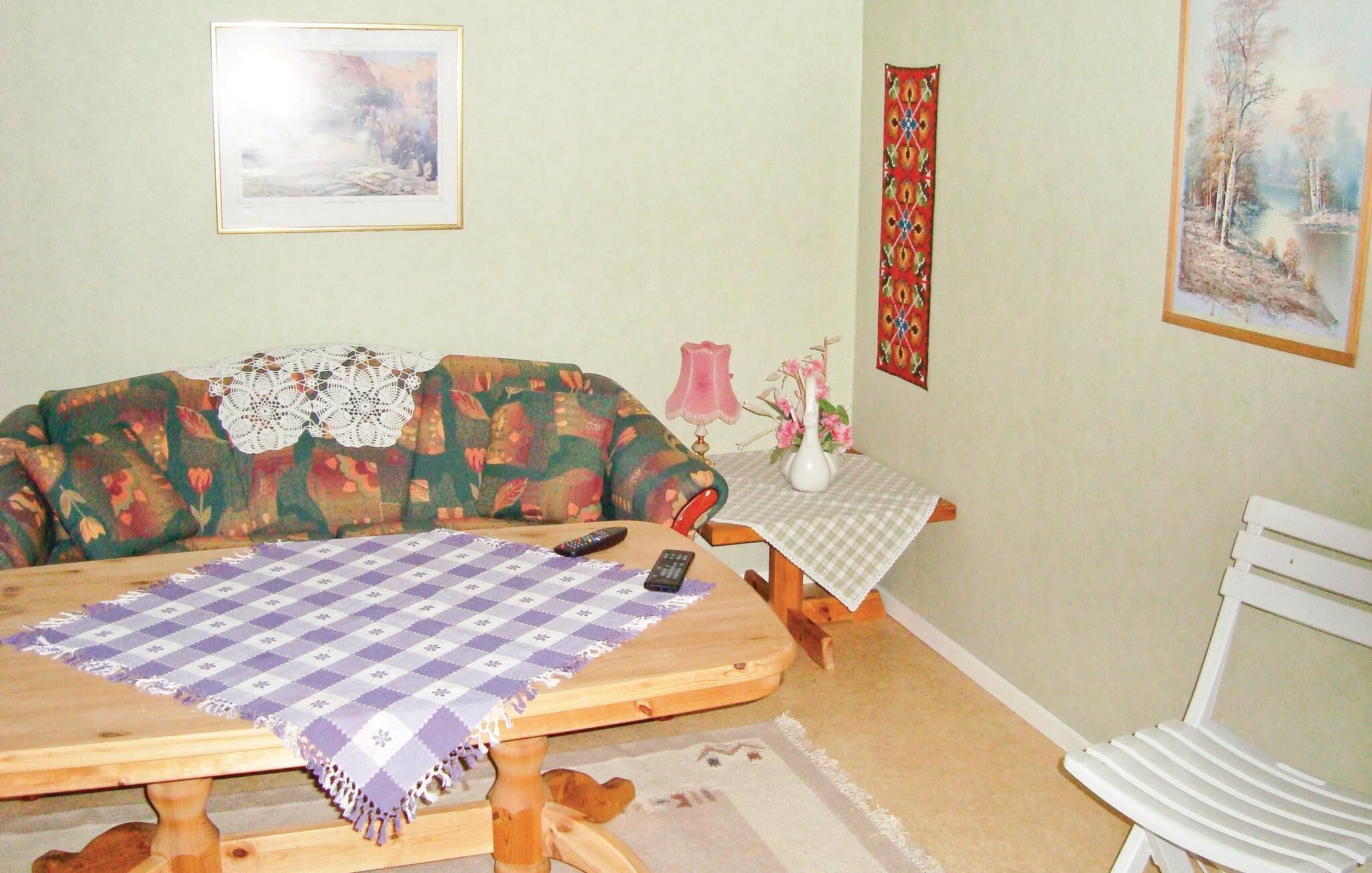 Una sala de estar acogedora con un sofá floral, una mesa de centro de madera con mantel a cuadros, una mesita con un jarrón de flores y un cuadro en la pared.