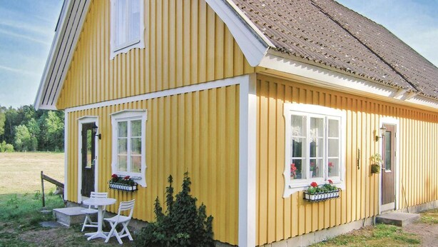 A yellow building with two windows and a door, a small table and chair outside, and a garden area. - 1 bedroom awesome home in Strömsnäsbruk (Strömsnäsbruk)