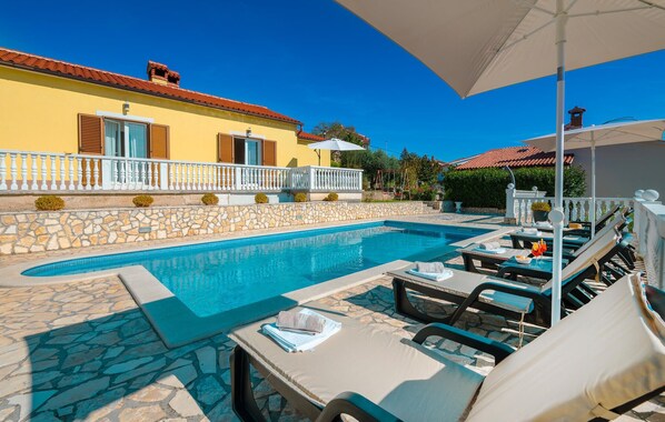 Ein Poolbereich mit Liegestühlen und Sonnenschirmen, eine Steinmauer und ein gelbes Gebäude mit Fensterläden.