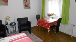 Une pièce comprenant une table à manger, des chaises, une petite table d’appoint avec un vase et une radio, ainsi qu’un lit recouvert d’une couverture à rayures.