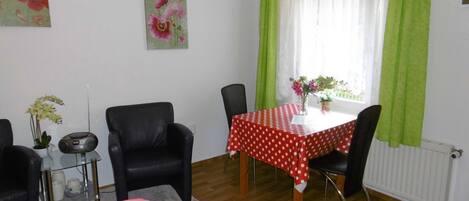 A room with a dining table, chairs, a small side table with a vase and a radio, and a bed with a striped blanket.