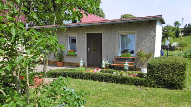Une petite maison avec un toit rouge, une porte en bois et une fenêtre. On y trouve des plantes en pot et un banc à l’extérieur.