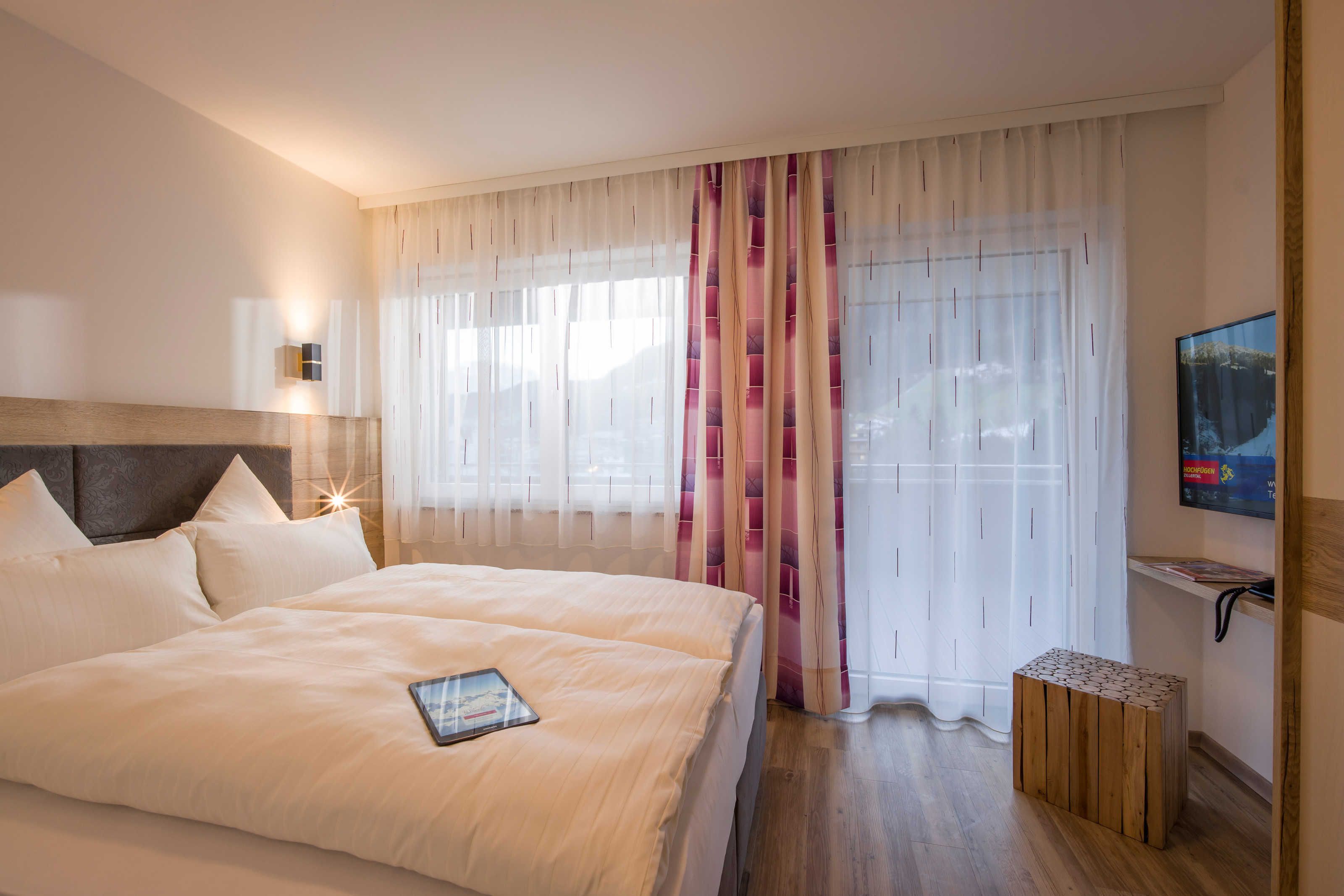 Hotelkamer met een groot bed, nachtkastje, tv aan de muur en een raam met dunne gordijnen.