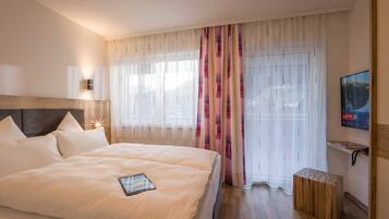 Hotelkamer met een groot bed, nachtkastje, tv aan de muur en een raam met dunne gordijnen.