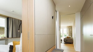 A modern hotel room with a wooden bench, a sofa with cushions, a bedside table, and a view of the hallway leading to the living area. - K6 from A&M - K6, Germany (Kampen (Sylt))
