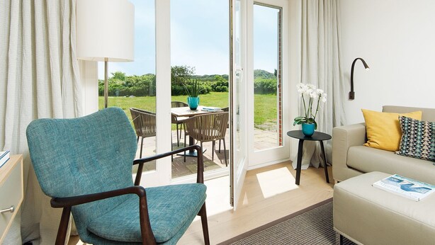 A modern living room with a teal armchair, a beige sofa with yellow and patterned cushions, a small side table with a blue vase and white flowers, and a large window with a view of a green landscape. - K6 from A&M - K6, Germany (Kampen (Sylt))