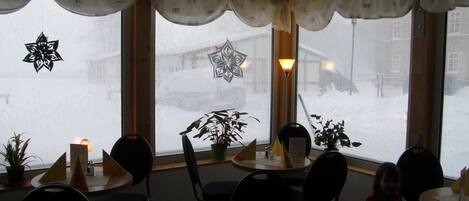 Un restaurante con mesas y sillas redondas, plantas en macetas y vistas a una calle nevada a través de las ventanas.