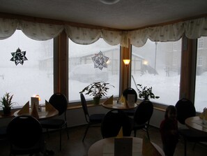 A restaurant with round tables and chairs, potted plants, and a view of a snowy street through the windows.