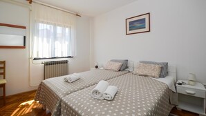 A hotel room with two beds, a window with curtains, a radiator, a bedside table with a lamp, and a framed picture on the wall. - Holiday apartment with terrace and air conditioning (Pula)