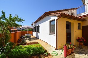 Situated in a residential neighborhood, this property features a compact single-story home with a flat roof and attached garage, surrounded by a well-maintained garden and outdoor seating area.