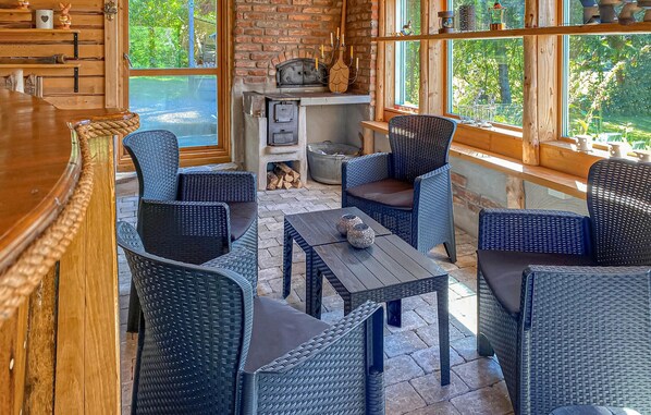 A cozy outdoor seating area with wicker furniture, a stone floor, and large windows offering a view of greenery.