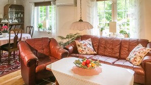 Ein gemütliches Wohnzimmer mit einer Ledersofa, einem Esstisch mit Obst-Dekoration und Blick auf Grün hinter großen Fenstern.