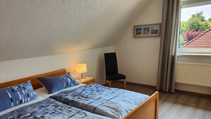 A bedroom with a wooden bed, a nightstand with a lamp, a chair, and a window with a view of trees and a building. - 101 2-room Spülm. (LTO,MNC)(BC.n8) - BUE - Haus Leißner (Büsum)