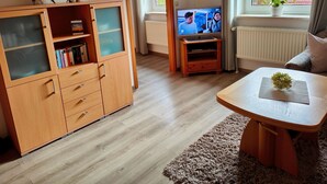 A cozy living room with a wooden cabinet, a television, a small table with a plant, and a view of the outside through a window. - 101 2-room Spülm. (LTO,MNC)(BC.n8) - BUE - Haus Leißner (Büsum)
