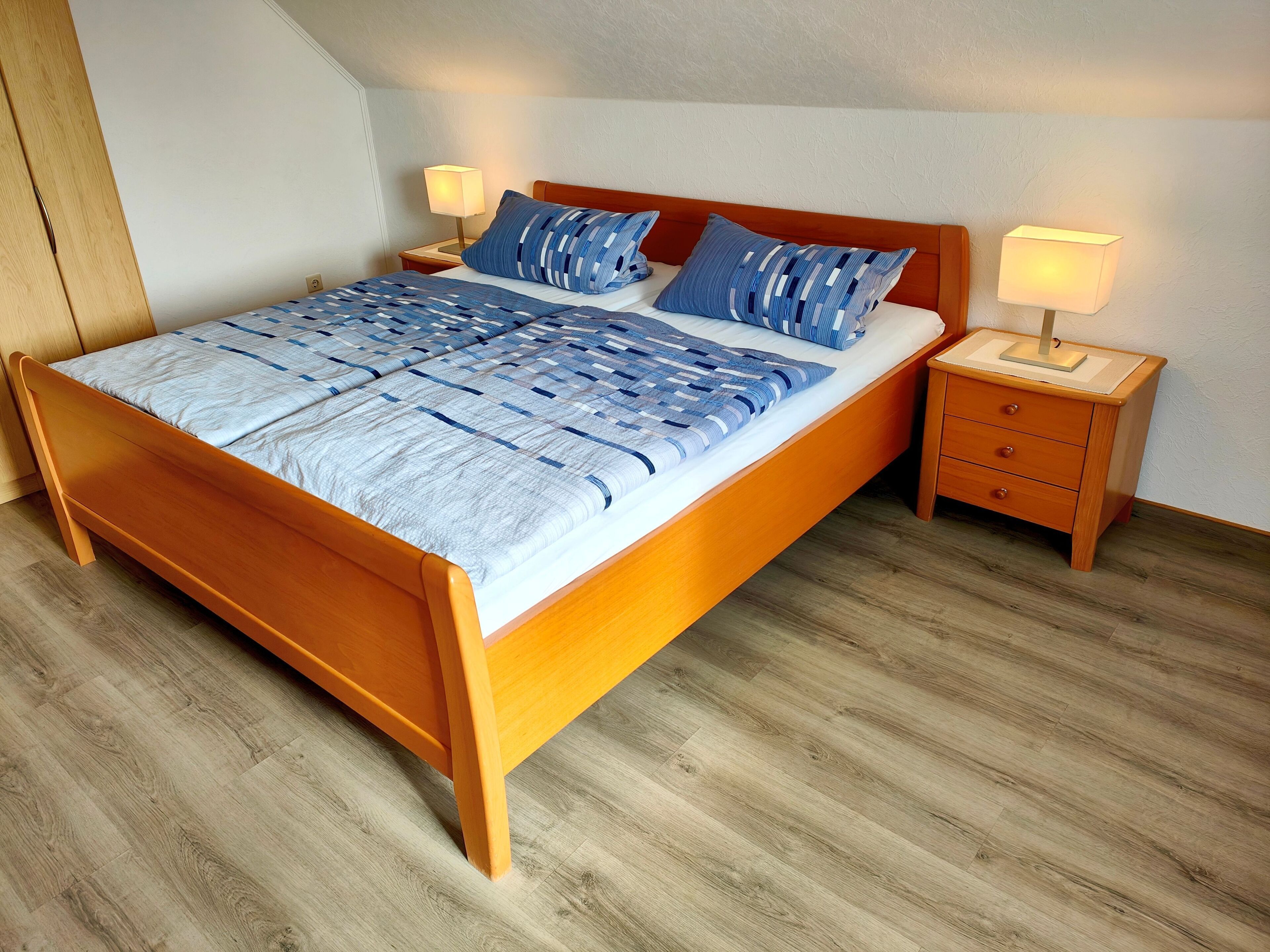 A wooden bed with a blue and white quilt, two bedside tables with lamps, and a wooden floor.
