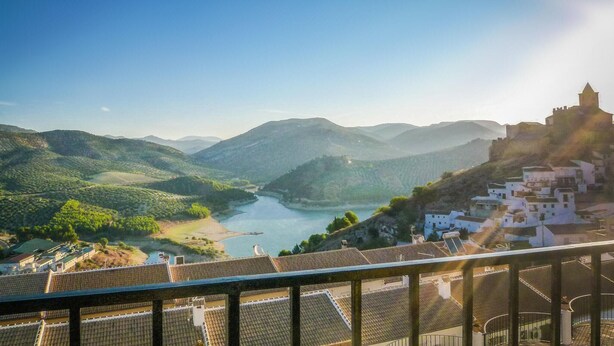 Une vue pittoresque d’une vallée traversée par une rivière, avec un château perché sur une colline, entouré d’une végétation luxuriante et de bâtiments blancs.