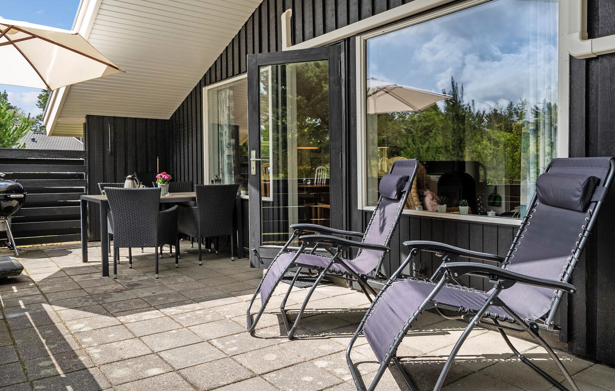 An outdoor patio with a table and chairs, a grill, and a large window overlooking a garden.