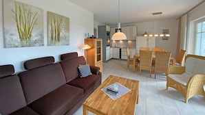 A modern living room with a sofa, a wooden coffee table, and a dining area with a table and chairs. There is a kitchen in the background with cabinets and a hanging light fixture. - MR: Villa Wilhelmine Whg. 02 Wilhelm with terrace - Villa Wilhelmine Whg. 02 (Sellin)