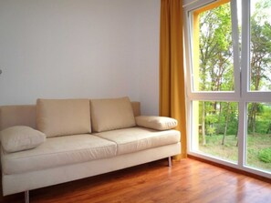 Un canapé beige dans une pièce dotée d’une grande fenêtre, d’un plancher en bois et offrant une vue sur la verdure à l’extérieur.
