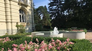 A garden with a fountain, pink flowers, and a building with classical architecture. - Residence Bleichröder App. Cosima, VS Sass (Heringsdorf)