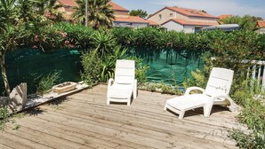 A wooden deck with two white lounge chairs, a small pond, and a backdrop of houses with red-tiled roofs. - Stunning home in Torreilles - Plage (Torreilles - Plage)